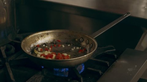 Focused professional chef cooking peppers and onions in a saucepan on a stovetop in industrial kitchen with soft lighting. Close up shot 4k RED camera.