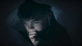 Night shot of teen boy with headphones using tablet pc under blanket. Happy child playing with cellphone or smartphone on a bed. Light reflection on face cute young boy. 