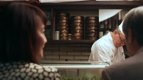 Happy Japanese couple sitting at a sushi bar watching the expert chef slice fish in small sushi bar with soft interior lighting. Medium shot on 4k RED camera.