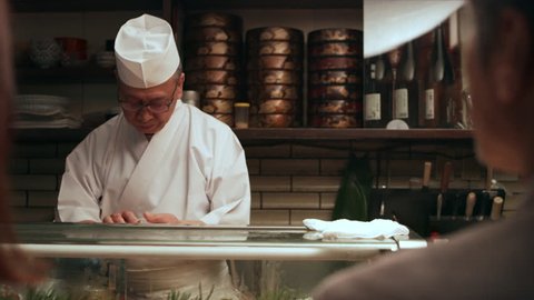 Expert chef prepares dinner for happy Japanese couple sitting at a bar counter getting ready to eat sushi in traditional sushi bar with soft interior lighting. Close up shot on 4k RED camera.