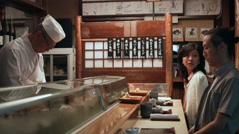 Timid Japanese couple on date drinking tea and watching while chef cooks in small sushi bar with soft interior lighting. Close up shot on 4k RED camera.