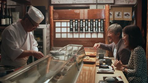 Smiling Japanese couple eating sushi and chatting with chef while he cooks in small sushi bar with soft interior lighting. Close up shot on 4k RED camera.