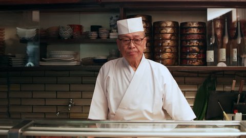 Portrait of veteran sushi chef with small smile standing behind a counter in his small traditional sushi bar with soft interior lighting. Medium shot on 4k RED camera.