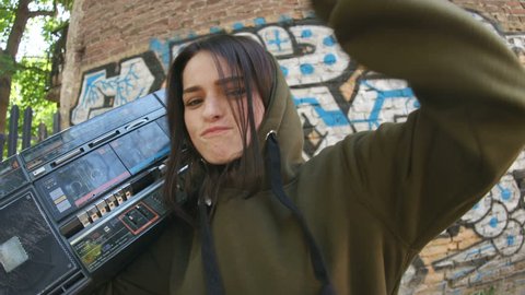 Cool stunning brunette with retro boombox looking into camera enjoying music, dancing. Portrait of cheerful girl in background of street art. Graffiti on wall. Outdoors.