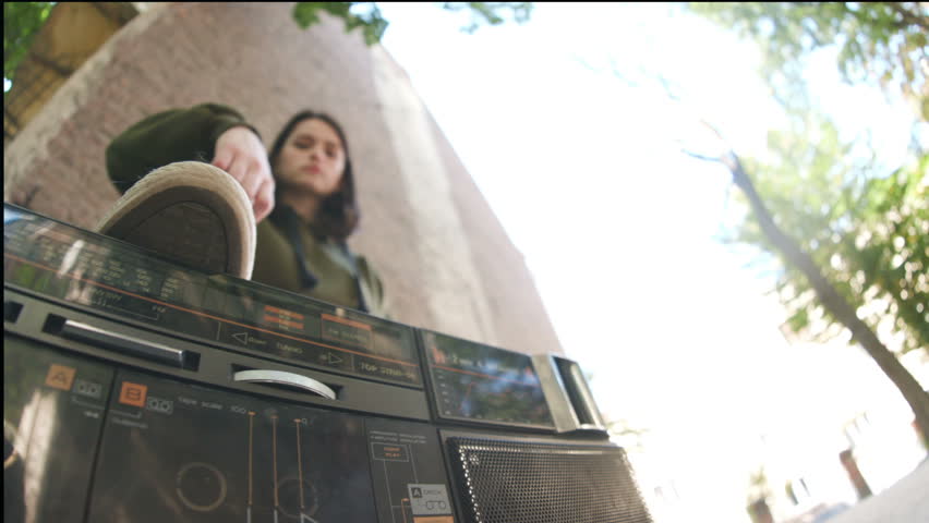 Cool young Caucasian person in sport clothes standing foot on portable stereo boombox. Good-looking student looking at camera. Daytime. View from below. Lifestyle.