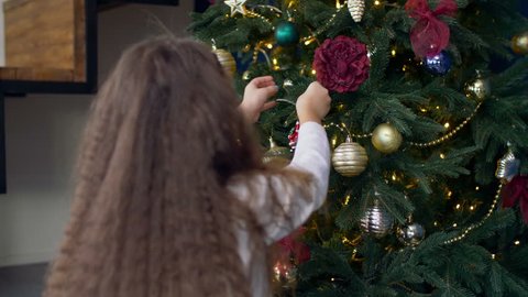 Rear view of little girl with long brown hair decorating Christmas tree with toys at home. Happy excited child in anticipation of Christmas and winter holidays, decorating Christmas tree.