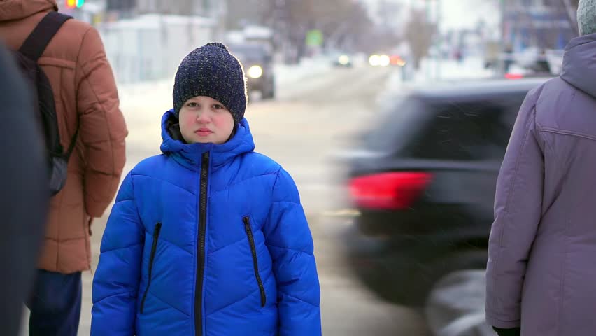 Boy in a blue down jacket standing on the street. Cars are riding in the background, the boy is watching.