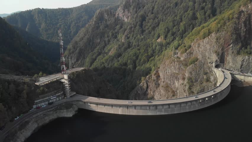 Drone footage - Drone flying over the dam heading to the mountains, where cars are riding.