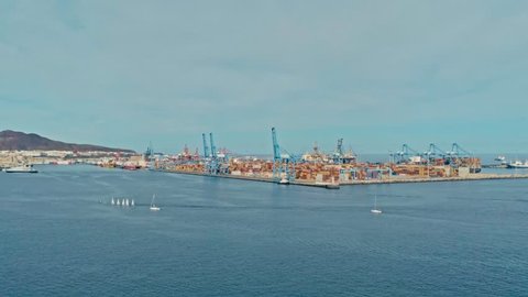 aerial drone footage of the harbor with numerous containers piled up next to cranes ans a small sailing dinghy race 
