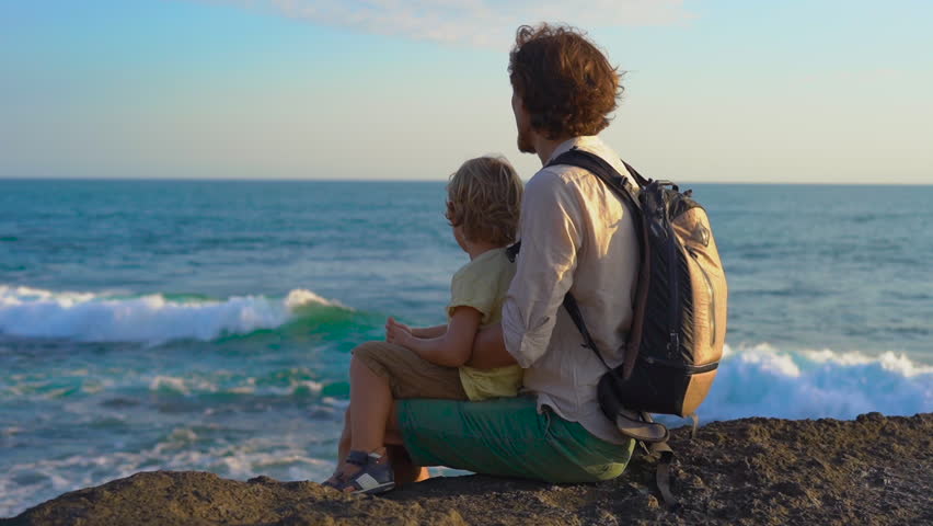 Slowmotion shot of a father and son sitting on a rock looking at an ocean waves nearby the Tanah Lot temple