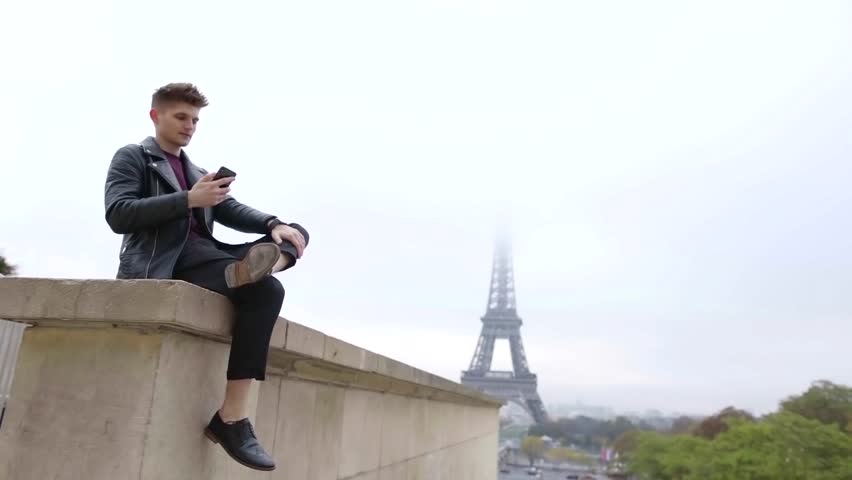 alone handsome man is sitting on stone wall in Paris in fall day, reading messages in mobile phone