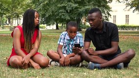 A black family sits on grass in a park, looks at a smartphone and talks about something