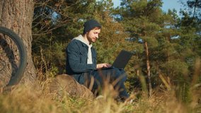 Active and mobile young man with laptop sitting in a clearing and networking in the country.