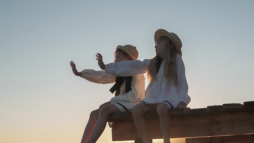 Happy girls in beautiful dresses are playing on the sea pier. In the background sea sunset. Wooden pier
