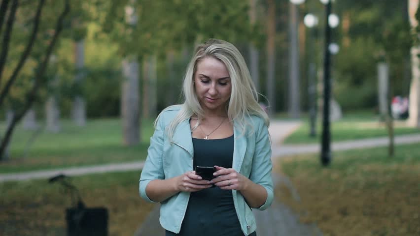 An attractive young woman reads and writes messages while walking in a summer park on a sunny day | Shutterstock HD Video #1019966983