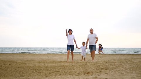 A happy family of three walking along the sandy sea beach, looking towards the sky, showing with a hand, in slow motion.