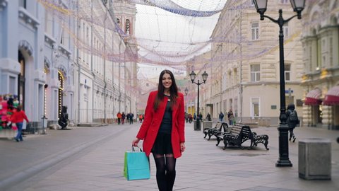 Fashionista long haired woman in red coat is walking outdoors in the street. She has shopping bags in her hands. 