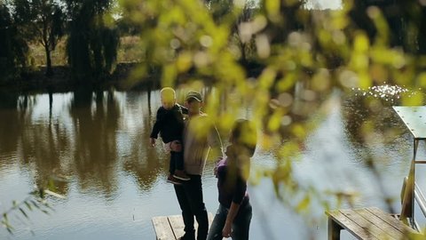 A young family of three people under a green tree on the shore of a lake in autumn park. Slow motion