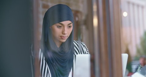 Young muslim woman wearing hijab sitting in modern cafe, working, searching or studying at her laptop, drinking morning coffee - modern muslim concept closeup 4k