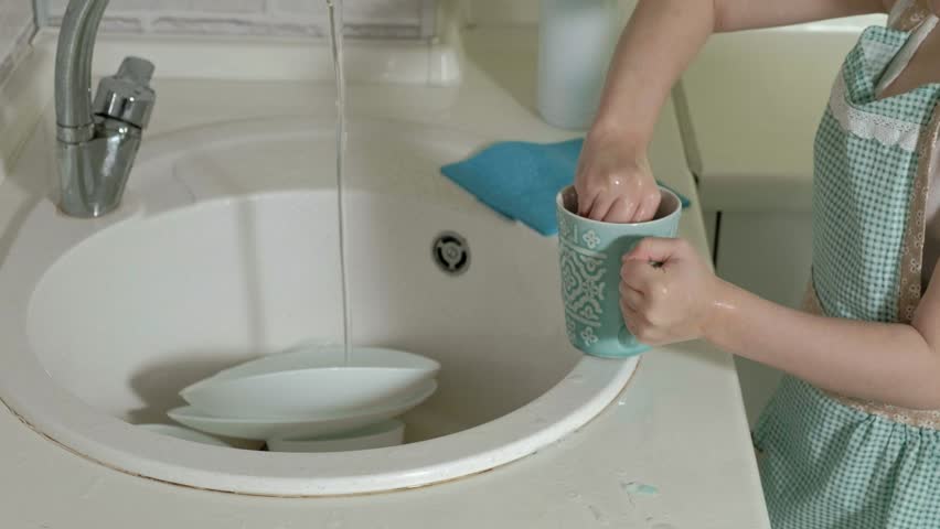 beautiful little girl in an apron is standing on a chair, in the bright kitchen, washes a cup with a sponge, helps parents 4k