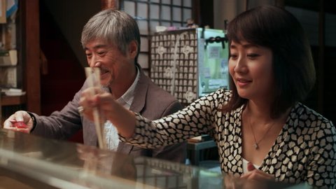 Hungry Japanese couple using chopsticks to eat sushi by the counter where the Master chef works in small sushi bar with soft interior lighting. Close up shot on 4k RED camera on a gimbal.