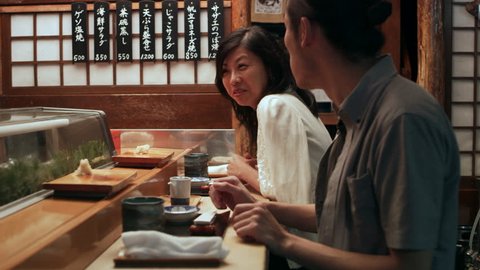 Hungry Japanese couple scarf down sushi while talking with the head chef in small sushi bar with soft interior lighting. Medium close up shot on 4k RED camera.