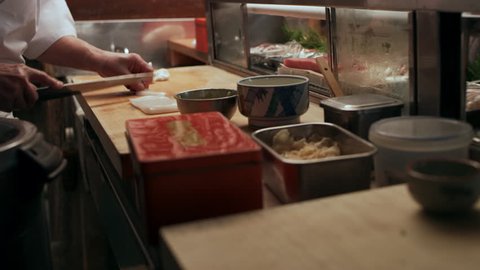 Sushi chef slicing pieces of Red Snapper and Tuna fish and rolling a ball of rice to make sushi for two customers in small sushi bar with soft interior lighting. Close up shot on 4k RED camera.