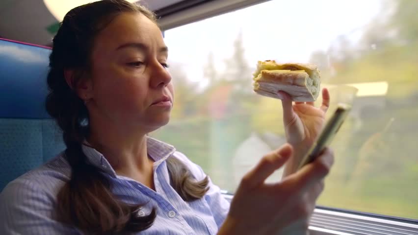 Woman eating sandwich and using smartphone on a train, woman traveling.