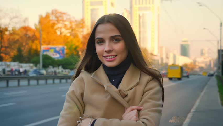 Gorgeous female of caucasian appearance is standing calmly and hidning behing her coat in aoutumn sunny day near road, cars in motion on backgound