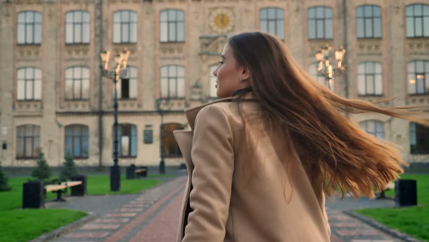 Charming caucasian female with long brown hair is walking ahead from camera and looking aside in motion, urban illustration outdoor, modern person in city