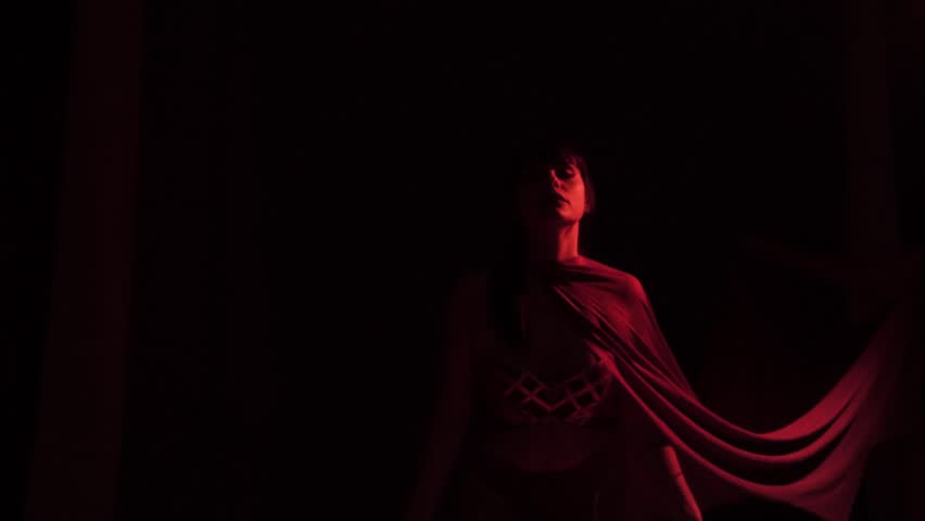 charming brunette with bangs and a ponytail stands motionless with head lifted and eyes closed, dressed in a skirt and a top with gold stripes, her cape easily flies forward because of the wind