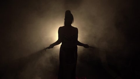 mysterious dark silhouette shadow of power creepy scary horror warrior woman stands alone in room smoke fog bright light behind her back, crosses waves her swords, two katana. Art Authentic lifestyle