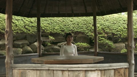 Portrait of a Japanese woman sitting at a table in a gazebo in a rocky and green garden in Japan with soft natural lighting. Medium shot on 4k RED camera.