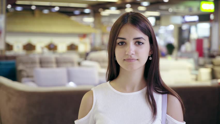 Portrait of a nice young brunet woman with long loose hair standing and smiling in a modern cafe of a modern mall with many sections in summer