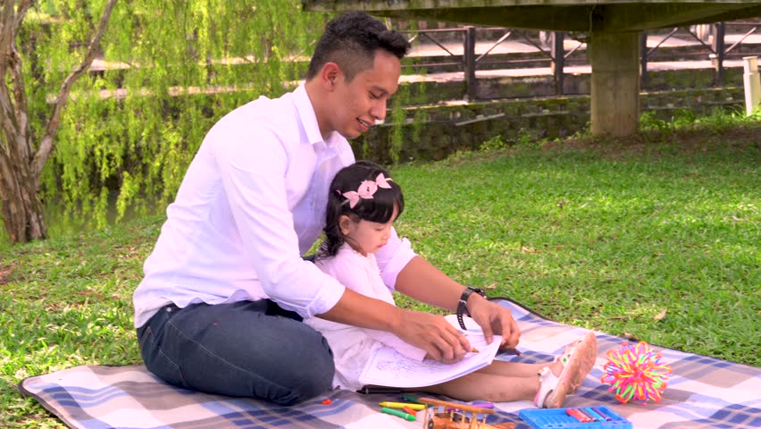 Young man teaching his daughter on coloring book with nice green park background. Royalty-Free Stock Footage #1020063763