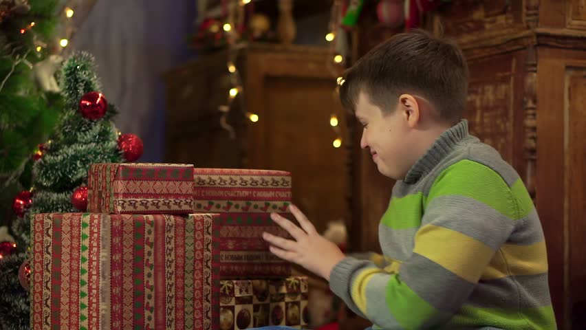 Boy teenager shakes and checks for Christmas presents. Gifts stacked under the tree