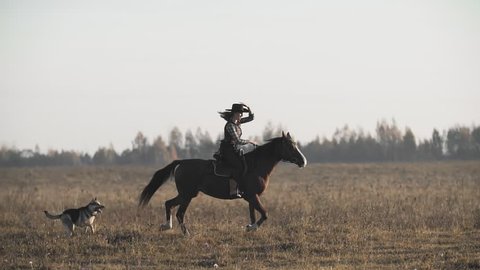 Beautiful woman riding a horse in background sunrise in field. Young cowgirl at brown horse in slow motion outdoors with happy dog