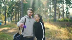 Sportive girlfriend and boyfriend are making online video call using smartphone standing in park together looking at device showing thumbs-up, waving hand and laughing.