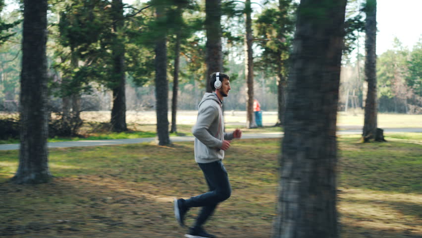 Dolly shot of bearded young man in tracksuit and sneakers jogging in park and listening to music through modern headphones. Sports and millennials concept.