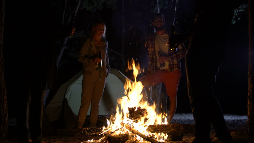 Smiling campers standing around bonfire with beer bottles, having great time