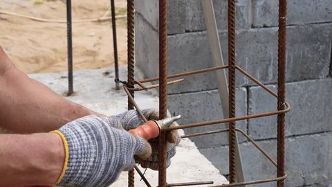 Hands of the builder in working dirty gloves tie the steel wire on the rebar with the help of a tool, creating a strong frame for reinforced concrete structures at the construction site of the house