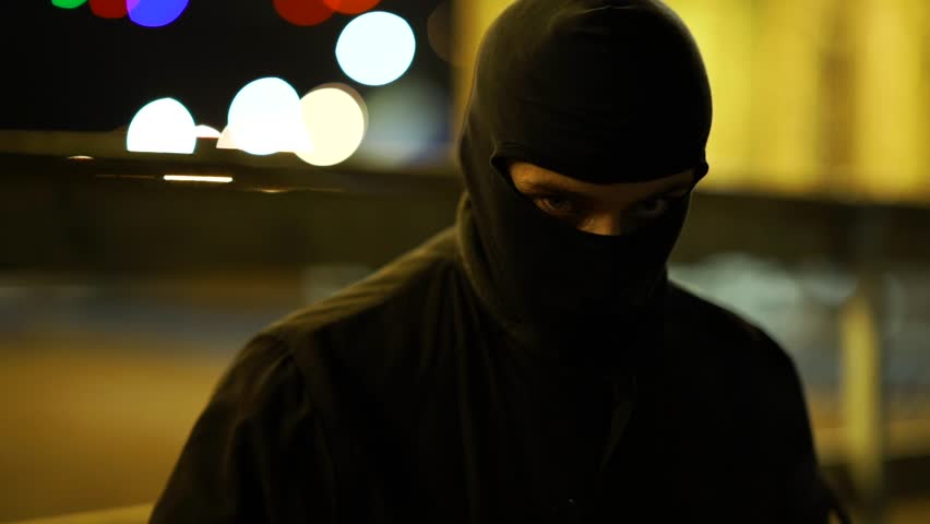 The thug looks at the camera. A man dressed in dark clothes and a balaclava goes with a bat in an abandoned park. There is an early winter outside and garlands hang around. | Shutterstock HD Video #1020091534