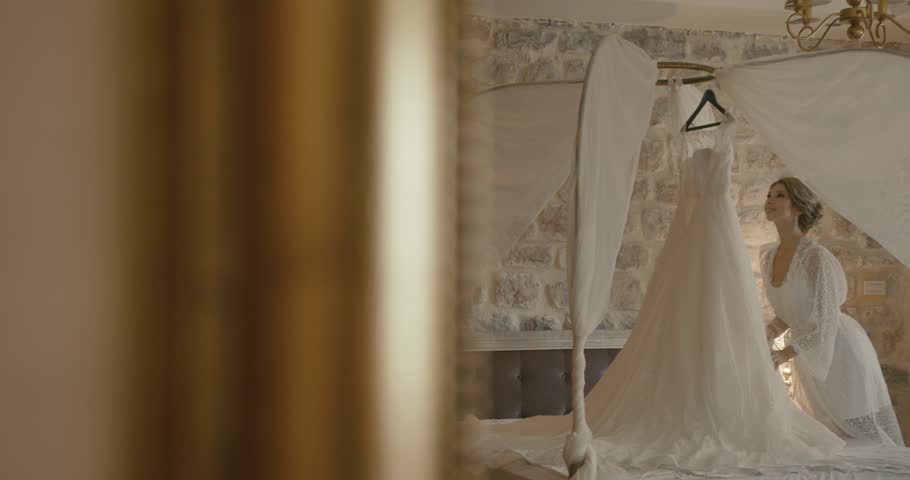 Wedding at Montenegro. Traditional European ceremony. Morning before the reception. 4k. Bride in delicate silk robe admires her wedding dress standing in a hotel room