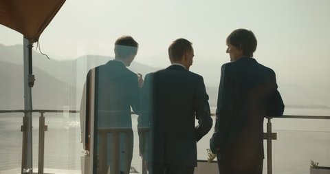 Wedding at Montenegro. Traditional European ceremony. Morning before the reception. 4k. Business men. Three men in blue suits drink whisky standing on the balcony with great mountain view