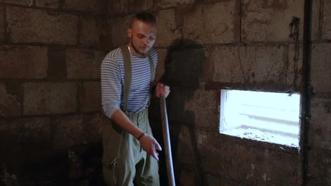The man shovel litter into a mound, he is holding a shovel. Medium shot, natural light, indoors, in a village on a farm, in a pigsty, young guy doing a kidney-buster