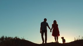 couple silhouette walking. slow motion video. holding hand toward the sun outdoors at sunset dramatic sky background and dog. young happy couple go on nature sunset sun light. man and girl silhouette