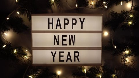 Happy New Year text displayed on a lightbox on the table with Christmas lights and fir branches. Night. New Year concept.