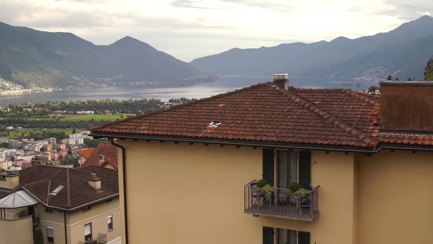 Medium Shot. Slow pan across homes in the mountainous region of Orselina, Switzerland with the city of Locarno far below. The Swiss Alps, on the shores of Lake Maggiore are in the distance.