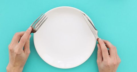 Female hands with cutlery and empty plate on turquoise background. Top view of nervous hungry female hands at dinner table holding fork and a knife above empty flat white plate