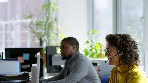 Tilt down of concentrated African businessman working on computer at desk while Caucasian woman talking with colleague in modern open space office
